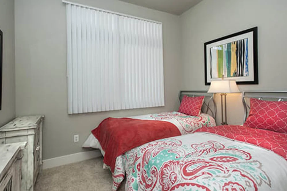 Twin-bedroom with two patterned twin beds, bedside lamp, dresser, and vertical blinds